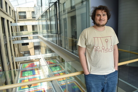 Thomas Bergamaschi, wearing a T-shirt that says "physics" and showing a modified Newton's cradle, leans on a railing overlooking an indoor courtyard with a colorful floor