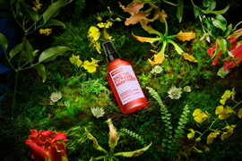 An orange spray bottle that has the text “Palmless” and its slogan on the label lays atop a grassy surface surrounded by tropic, colorful flowers.