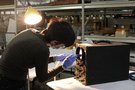 In a museum storage area, a side view of Florencia Pierri cleaning an electronic device that looks like an oscilloscope, which sits on a table in front of her. Metal shelves with many boxes and items are in the background.