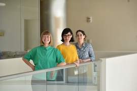 Taylor Baum, Josefina Correa Menendez, and Karla Alejandra Montejo stand in a line and smile along a glass railing in a corridor of MIT Building 46.