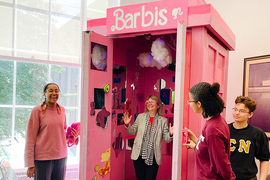 Sally Kornbluth is standing in a pink Barbie-themed tardis labeled "Barbis," wearing pink 80s sunglasses and holding up jazz hands. One student stands at left and two students stand at right. Everyone in the scene is laughing.