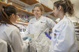 Nicole De Nisco and two others chat at a biology lab bench. All three are wearing white lab coats and hair in ponytails.