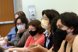 Four college students wearing masks sit at a table. One is gesturing with her hands.