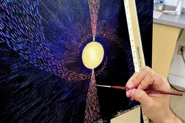 Hand holding a paintbrush, painting on a canvas showing what appears to be a bright yellow planet and pink and blue dots representing dust on a black background