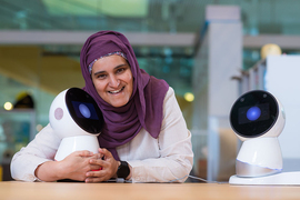 Sharifa Alghowinem, who wears a head scarf, leans on a table, hugging a Jibo personal robot, which looks like a sphere with a large eye.