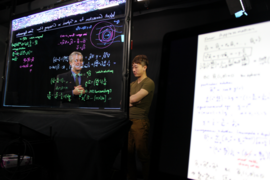 Martin Bazant and Joey Gu are seen through a clear writing board covered with mathematical equations.