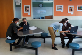 Four people share two laptops at separate tables in a tidy cinderblock room. 