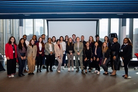 A group of about 25 people pose together against a large window with Boston’s skyline in the background