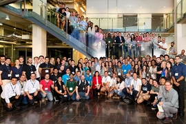 Hundreds of MIT Bootcamps alumni pose in a large atrium, near stairs.