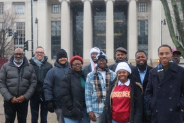 Eleven people, mostly with various shades of brown skin, pose in winter jackets before the columns of MIT's front entrance. It’s snowing.