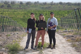 Three women, researchers from the GEAR Lab, stand on a dirt road in a field in Jordan holding laptops.