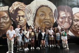 21 people pose in front of a mural depicting faces of five individuals of color