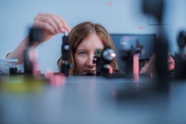 Kristina Monakhova, crouching, peers along the surface of a table of experiments with cameras and microscopes. Only her head from the mouth up is visible.