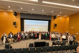 About 65 people pose standing in a semicircle on a large auditorium stage, with the backs of empty seats in the foreground and a video screen behind them.