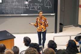 Sossina Haile, a Black woman with short hair, stands in front of a blackboard covered in equations, speaking to an audience