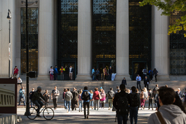 Pedestrians cross the street in front of MIT's Massachusetts Avenue entrance, with stairs leading up to large neoclassical columns.