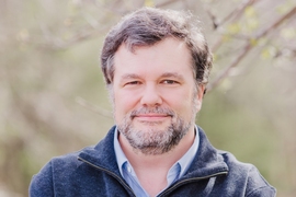 A bearded man outside in an open-collar fleece pullover regards the camera with a faint smile