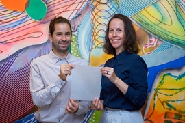 A man and a woman , stand in front of a colorful background. They're holding a piece of translucent, paper-like biomaterial packaging.