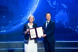 Susan Solomon and Tran Hong Ha pose on a stage. They are both holding a folio open for the audience to see, and Solomon is holding a gold trophy.