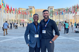 Runako Gentles and Shiv Bhakta pose together in front of the entrance to Expo City Dubai, a sand-colored building featuring a domed central structure. Flags of many nations fly in front of the building.