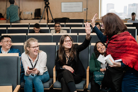 In a lecture hall, a person with a red shawl high-fives a seated person while several others look on