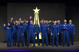 12 astronauts in blue jumpsuits appear on stage. Several are clapping or waving at the crowd.