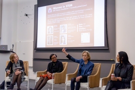 Four women sit on a stage, one with a raised fist, in front of a projected slide headlined "Women in STEM."