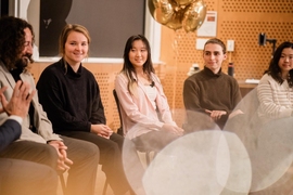 Five people seated in a row in front of an audience smile and listen intently to someone out of the frame.