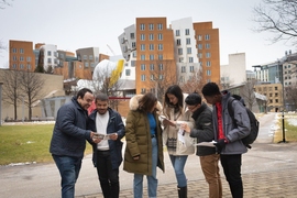 Six students wearing winter jackets consult printed materials and talk with each other in front of the Stata Center, a building of brick and steel with unusual angles