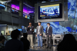 Three students stand on a stage with a large monitor behind them displaying a group of people and the words "Thank you."