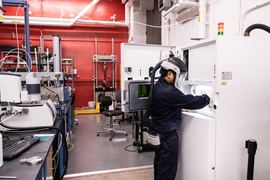 A student wearing protective gear including a full helmet and face shield with air supply works at a refrigerator-sized machine