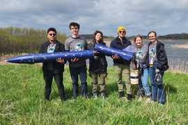 Six people dressed in team T-shirts and jackets pose in front of a pond. They're holding a large blue rocket with gold star designs.