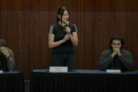 Claire Wang stands on stage with a microphone and two other competitors are seated.