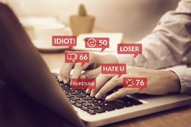 Hands typing on a laptop emitting word bubbles with hateful comments like "IDIOT!" and "LOSER.”