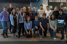 17 people pose in two rows for a group photo. One holds up a laptop screen to include half-a-dozen remote participants.