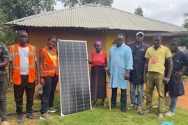 8 people pose standing in front of a small building and showing a solar panel. One person is mostly outside of the frame.