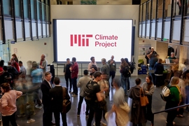 A crowd of people mill about an atrium in front of a large screen with the MIT Climate Project logo.