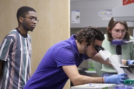 Two students look on as an instructor peers into a microscope