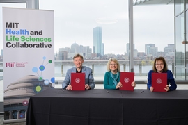 Vincent Roche, Sally Kornbluth, and Anne Klibanski sit side by side at a table, each holding up a red folder with the MIT seal 