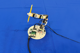 A small robotic arm is mounted on a circular white base, on a bright blue surface. It is holding a yellow pencil vertically and is connected to wires and electronic components, including a Raspberry Pi board.