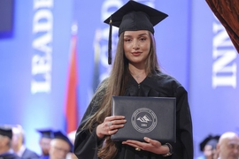 Woman with long brown hair wears a cap and gown and looks at the camera while holding her diploma
