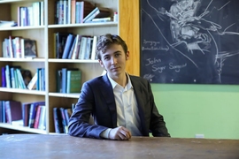 A student wearing a suit jacket and white dress shirt sits at a table in a lounge containing bookcases and a messily erased blackboard.