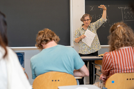 Leslie Kaelbling stands in front of a chalk board, holding one hand up in front of a group of students in chairs.