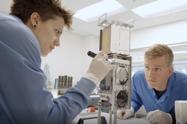 Two engineers inspect a small CubeSat (about the size of a loaf of bread) on a lab table. 