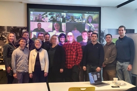 14 people stand around a large projection screen displaying at least a dozen other individuals on Zoom.