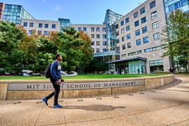 Student walks by the MIT Sloan School of Management sign in front of building E62 on a sunny day