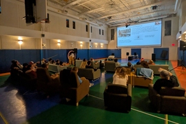 A couple dozen people sit in armchairs inside a gymnasium while watching a presentation projected on a large screen