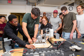 A diverse group of students huddles around a lathe on a classroom workbench. One manipulates the lathe while two hold it down.