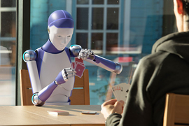 Photorealistic illustration of a humanoid robot seated at a table and playing cards with a human whose back is to the viewer
