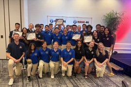 A group photo of about 30 RASC-AL winners, all wearing their team uniforms, with four students holding their award certificates. Everyone is smiling and some hold up other props.